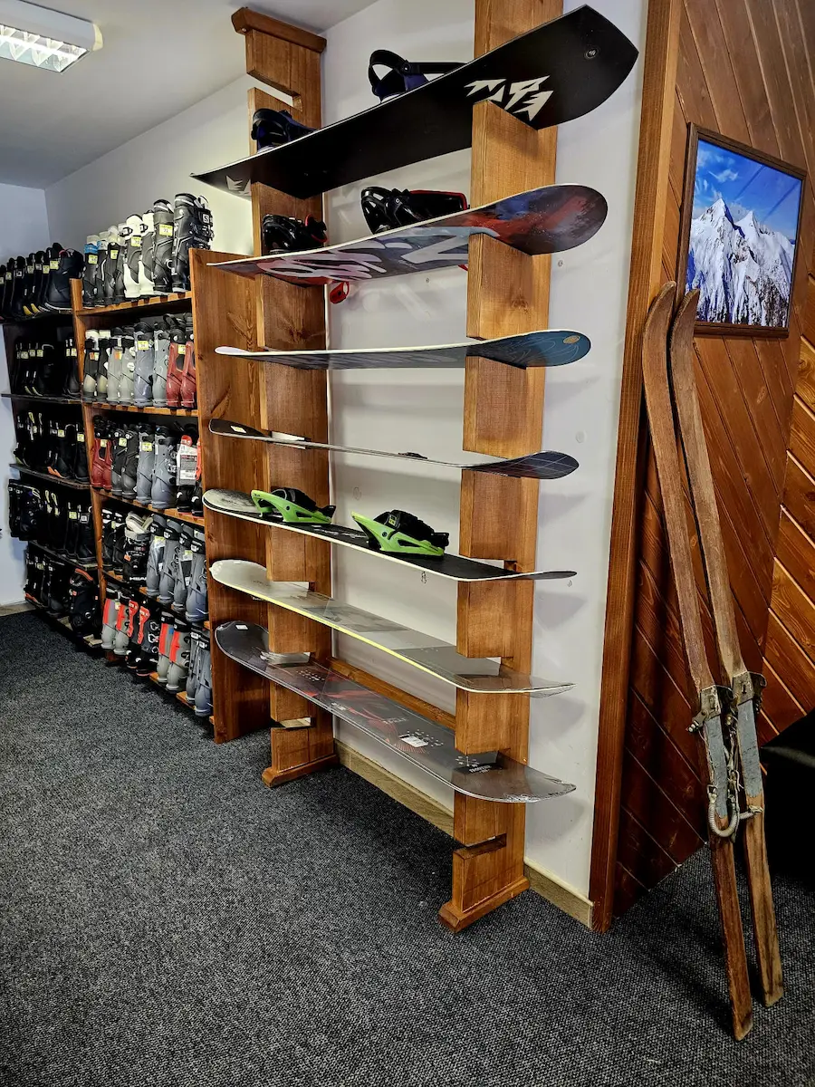 Interior of a BSR ski rental shop featuring a wooden wall-mounted rack holding multiple snowboards, a background shelf stacked with assorted ski boots, and vintage wooden skis leaning against a wood-paneled wall.
