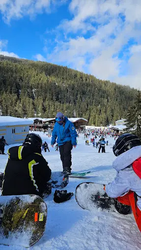 Group of snowboarders getting ready on a snowy slope near a busy mountain lodge with forested hills and blue sky in the background.