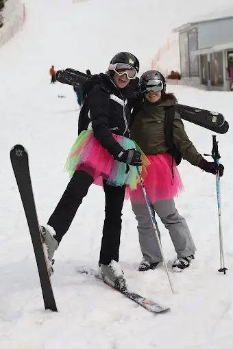 Two smiling skiers in helmets and goggles wearing bright tutus over ski gear, posing with skis on a Bansko snowy mountain slope.