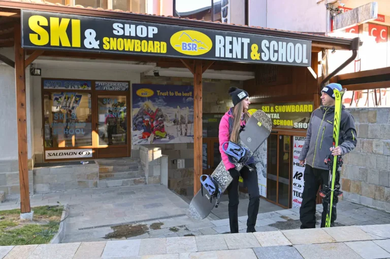 Couple standing outside a ski and snowboard rental and school shop, woman holding a snowboard and man carrying skis beneath a large “SKI & SNOWBOARD RENT & SCHOOL” BSR sign.
