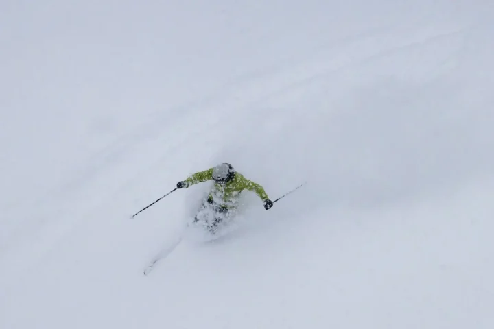 A skier in a green jacket carves through deep powder snow, creating a spray, seen from above in a snowy Basnko mountain landscape.