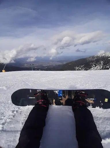 Snowboarder's legs and board on snowy slope overlooking mountains and cloudy sky with scenic valley in the distance.