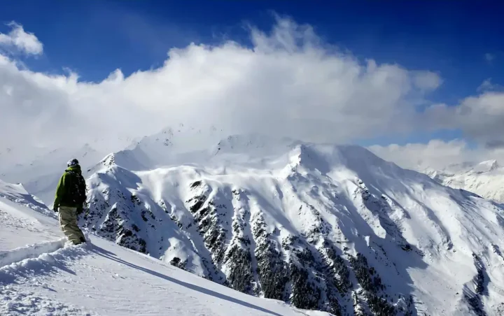 A snowboarder in green jacket and beige pants stands on a snowy ridge edge, gazing at expansive snow-covered mountain peaks beneath a partly cloudy blue sky.