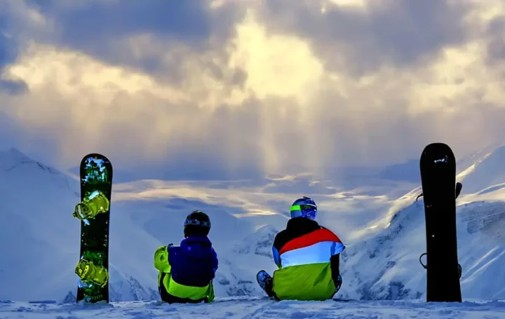 Two snowboarders in colorful jackets sit on a snowy ridge beside their upright boards, gazing at a vast sunlit mountain valley under a dramatic cloudy sky.